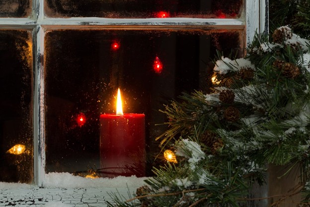 Red candle burning on a snowy window ledge with evergreen branches and soft glowing lights outside.